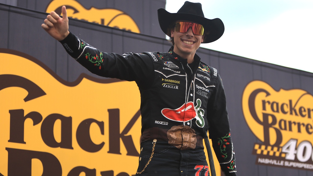 Home 1 A race car driver in a cowboy hat and sunglasses gives a thumbs up while standing in front of large Cracker Barrel 400 signs at a racetrack.