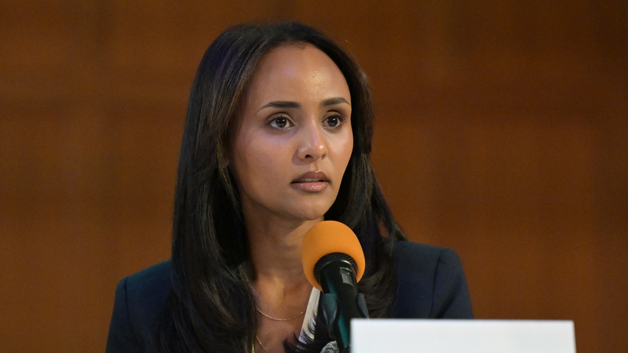 A woman with long dark hair speaks into a microphone with an orange windscreen, wearing a dark blazer, sitting in front of a blurred brown background.