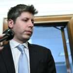 OpenAI CEO Sam Altman is surrounded by microphones, standing in front of an office doorway, appearing to speak or prepare to speak to reporters.