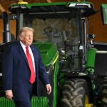 Trump walks past a large green tractor indoors, while people nearby watch and take photos.