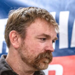Democrat Graham Platner looks down, with a microphone beside him and his campaign banner partially seen in the background.