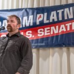 A man stands at a microphone with one hand raised, speaking in front of a campaign banner that reads Graham Platner U.S. Senate against a backdrop of white curtains.