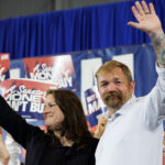 Graham Platner, with his wife by his side, wave to supporters with a campaign banner in the background.