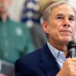 An older man in a blue suit holds a microphone, looking thoughtful. Two blurred people and a U.S. flag appear in the background.