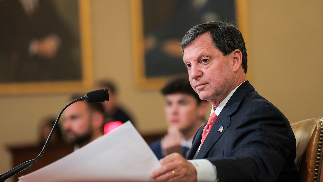 A man in a suit holds papers and sits at a desk with a microphone, looking to the side. Two blurred people and framed portraits are visible in the background.