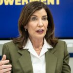 A woman with shoulder-length brown hair wearing an olive blazer and white blouse gestures while speaking, seated at a table with a water bottle. A blurred blue and yellow sign is behind her.
