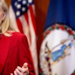 A woman in a red blazer speaks at a podium. Behind her are American flags and the Virginia state flag.
