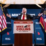 A man stands at a podium labeled Mike Collins U.S. Senate, arms outstretched, with American and Georgia state flags behind him and campaign signs in the background.