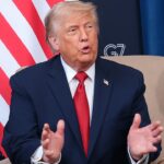 Trump in a dark suit and red tie sits in a beige chair, gesturing with his hands. Behind him are a partial U.S. flag and banners with G7 France 2019 and the U.S. State Department seal.