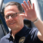 Gov. Josh Shapiro wearing a dark polo shirt waves, while another man stands behind him.