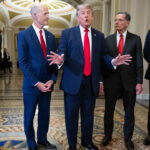 Five men in suits stand in a hallway with ornate flooring and archways. The man in the center gestures with both hands while discussing the Save America Act, as the others watch or look on seriously.