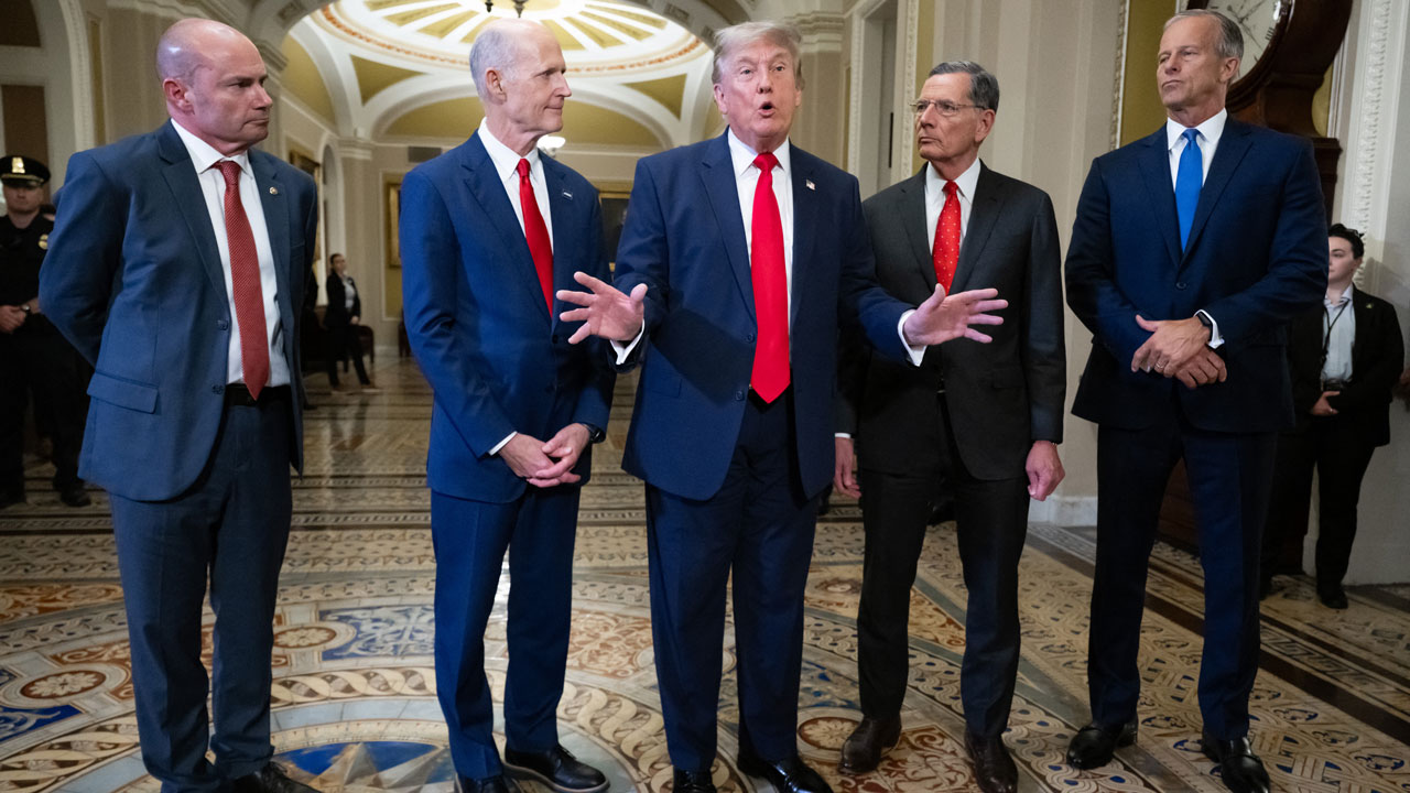 Five men in suits stand in a hallway with ornate flooring and archways. The man in the center gestures with both hands while discussing the Save America Act, as the others watch or look on seriously.