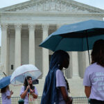 A group of young women stand around the Supreme Court with umbrellas.