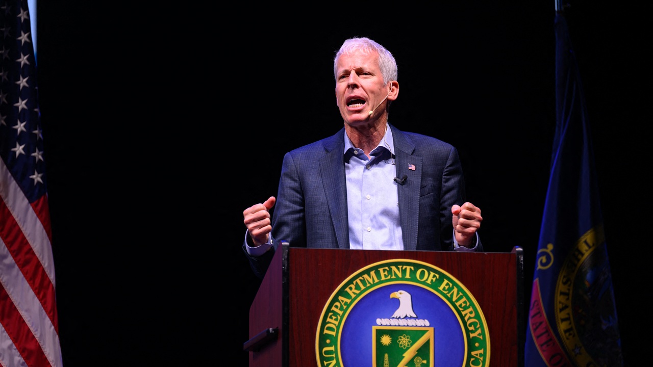A man speaks passionately at a podium with the U.S. Department of Energy seal, flanked by the American flag and another flag, against a dark background.