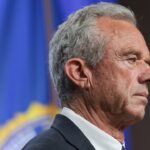 A man with gray hair in a suit stands in profile against a blue background with a blurred emblem or flag.