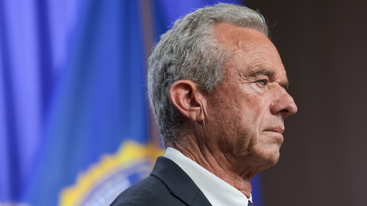 A man with gray hair in a suit stands in profile against a blue background with a blurred emblem or flag.