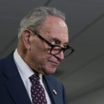 Chuck Schumer in a suit and patterned tie looks over the top of his glasses with a serious expression, standing indoors under fluorescent lights.