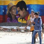 Four people walk past rubble in front of a mural featuring two people, a woman in a hat and a man in yellow, with a Venezuelan flag backdrop.