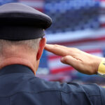 A police officer with a security wristband salutes in front of a large, blurred American flag backdrop, proudly honoring America.