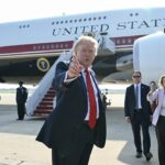 A man in a suit gestures while standing in front of a large plane labeled United States of America, surrounded by security personnel and reporters on an airport tarmac.