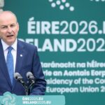 A man in a suit speaks at a podium with microphones. Behind him is a sign displaying ÉIRE2026.EU IRELAND2026.EU and related text about Ireland’s EU Council presidency in 2026.