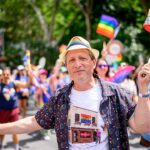 Brad Lander smiling and wearing a rainbow hat and patterned shirt stands at a Pride parade, surrounded by people waving rainbow flags and celebrating. Trees and buildings are visible in the background.