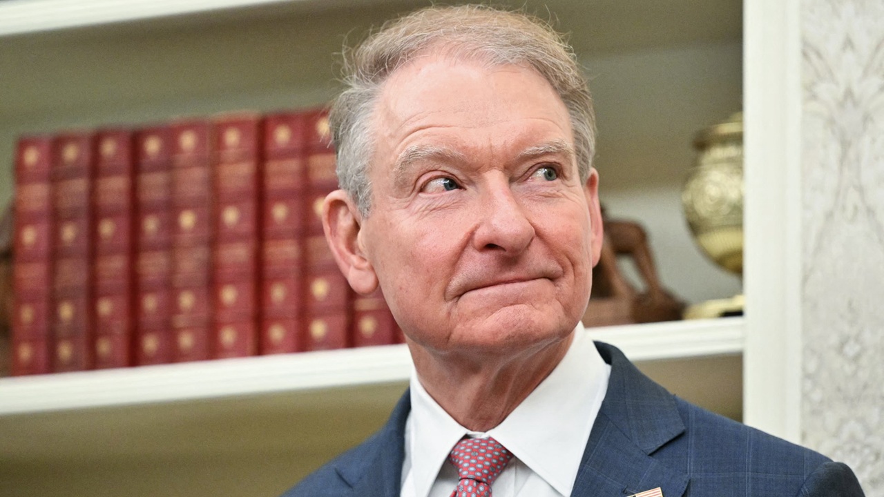An older man in a suit and tie stands indoors, looking to his left. Behind him are shelves with red books and a decorative vase.