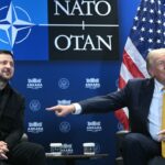 Two men sit beside each other in front of NATO, US, and Ukrainian flags. One man smiles with hands clasped while the other points to the side, both seated at a table with two cups.