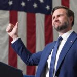 JD Vance Is Right: American Homelessness Is ‘Third World’ 1 A man in a blue suit gives a thumbs up while standing at a podium, with a large American flag hanging behind him.