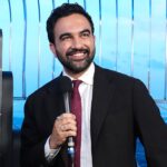 Zohran Mamdani in a suit and tie smiles while holding a microphone. He stands in front of a decorative background with gold designs and metal railings, with a cityscape visible behind him.