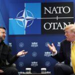 Two men in suits sit facing each other, gesturing while talking. Behind them are Ukrainian, NATO, and U.S. flags, along with a NATO OTAN logo.