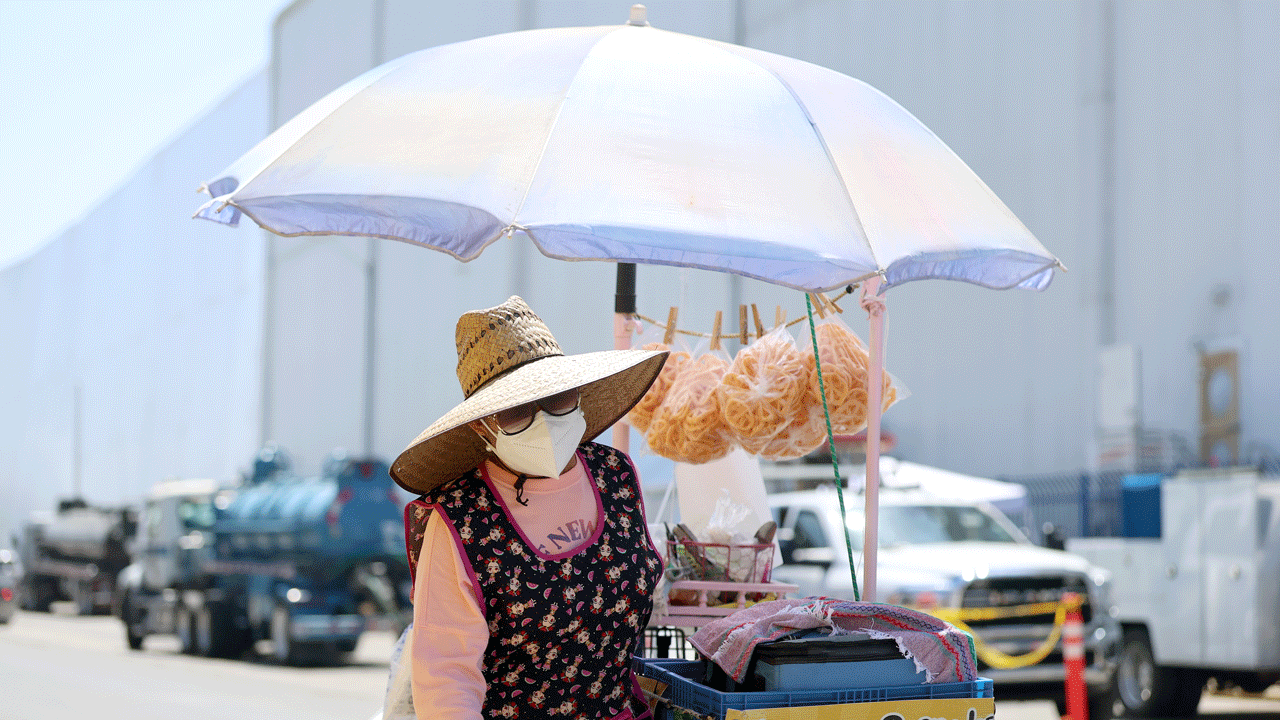 Home 3 A street vendor wearing a face mask and sunhat stands under a large umbrella, selling snacks displayed on a cart.