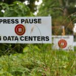 A sign among grass reads PRESS PAUSE ON DATA CENTERS with a red pause button symbol. More similar signs are seen in the background along a roadside.