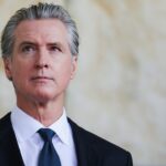 A man in a suit and tie with gray hair stands indoors, looking slightly upward with a neutral expression. The background is softly blurred in beige and cream tones.