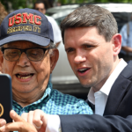 An older man wearing a USMC hat takes a selfie with a younger man in a suit. They are both smiling, surrounded by people outdoors on a sunny day.
