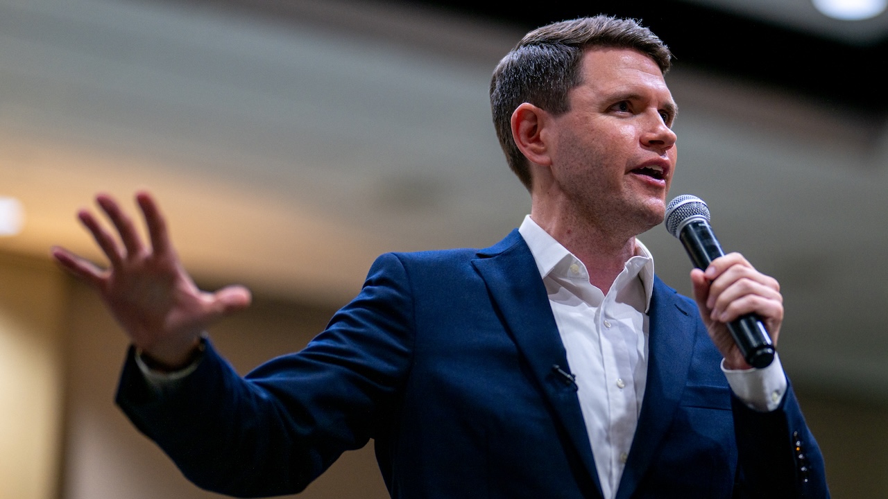 Home 4 James Talarico in a navy suit and white shirt speaks into a microphone, gesturing with one hand, in a brightly lit indoor setting.