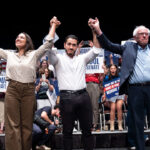 U.S. Rep. Alexandria Ocasio-Cortez (D-NY), Michigan Democratic candidate for Senate Abdul El-Sayed and U.S. Sen. Bernie Sanders (I-VT) holding up each other's arms at a rally.