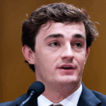 A young man, Nick Shirley, is in a suit speaks into a microphone, with a blurred brown background behind him.