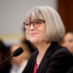 A woman with gray hair and glasses, wearing a black blazer and pearl necklace, speaks into a microphone at a formal event. Blurred audience members are seen in the background.