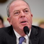 A middle-aged man in a suit and tie speaks into a microphone at what appears to be a formal hearing or meeting. He is looking slightly upward, with people blurred in the background.