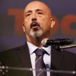 A bald man with a goatee in a suit and tie speaks at a podium with a microphone, gesturing with his hand. The blurred background features large orange text.