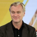 A man with light hair wearing a dark blazer and black shirt smiles while sitting in front of a colorful, blurred background with yellow and orange tones.