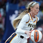A basketball player in a white Fever uniform dribbles a basketball during a game, with focus and intensity, as the crowd watches in the blurred background.