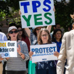 A group of people protest outdoors, holding signs. A man in sunglasses and a suit stands in the foreground.