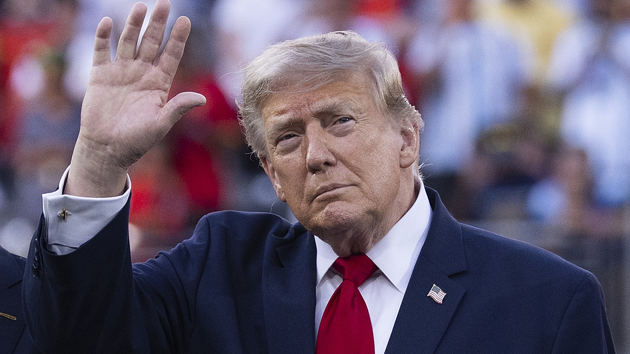 Home 5 A man in a navy suit and red tie raises his hand in a waving gesture. The background is blurred with people and various colors visible.