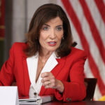 Gov. Hochul in a red blazer speaks at a table with an American flag in the background, discussing topics that touch on religion.