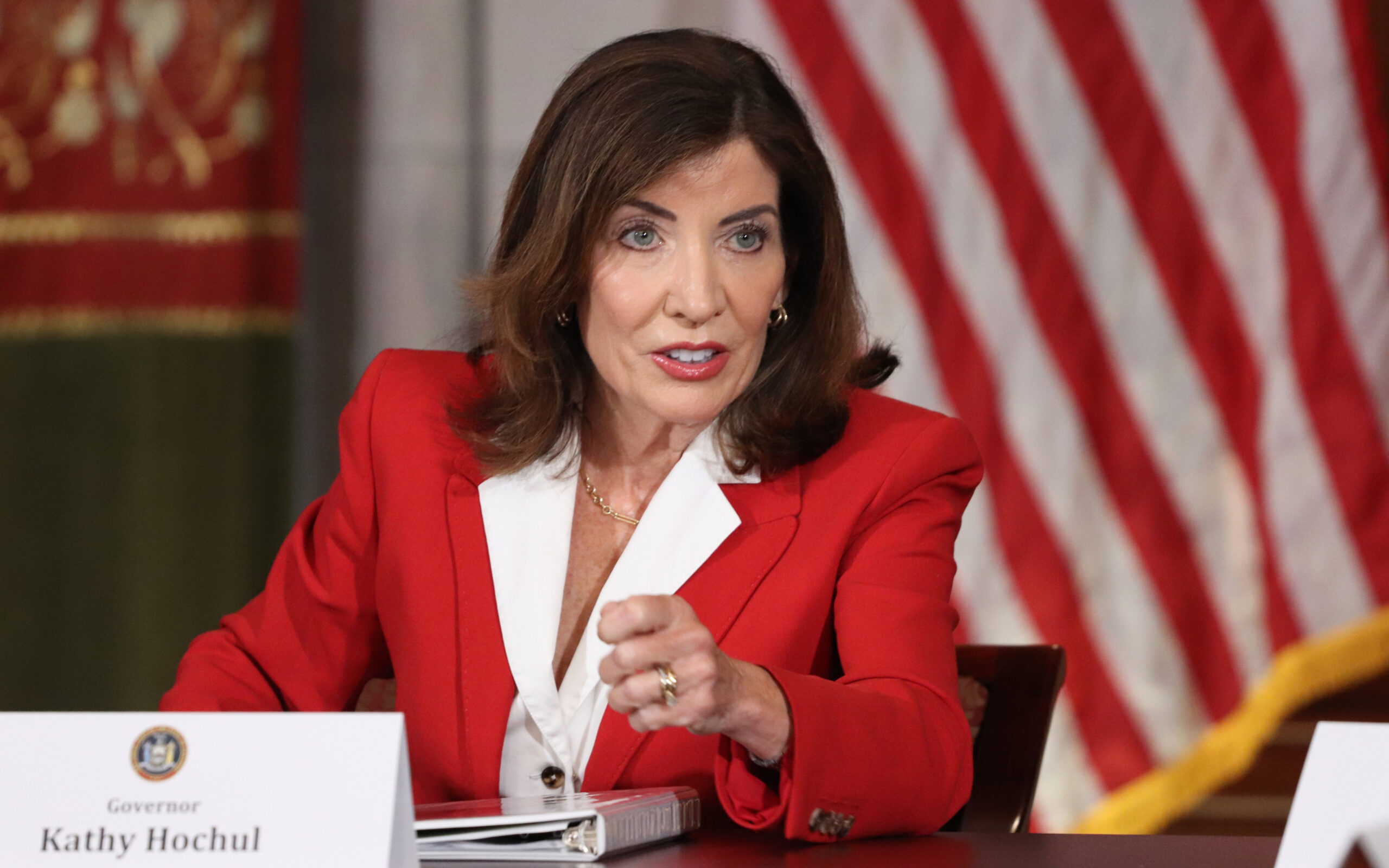 Home 1 Gov. Hochul in a red blazer speaks at a table with an American flag in the background, discussing topics that touch on religion.