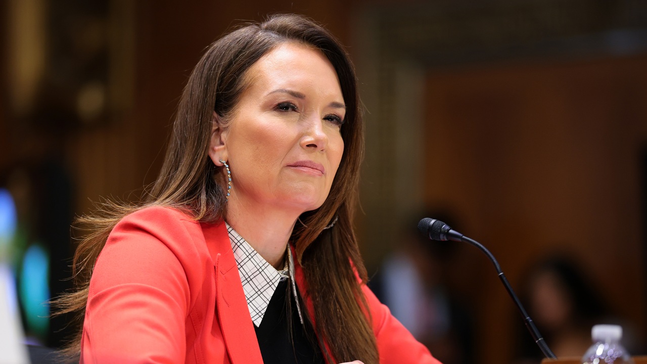 A woman with long brown hair in a red blazer sits at a table with a microphone, appearing to listen attentively during a formal meeting or hearing.