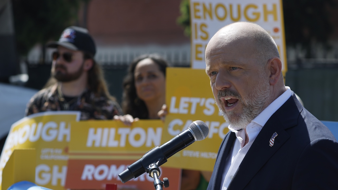 A man in a suit speaks into a microphone at an outdoor event. Behind him, two people hold yellow signs with messages like ENOUGH IS and LETS CA. The scene appears to be at a rally or protest.