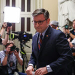 U.S. House Speaker Mike Johnson stands in front of reporters and photographers, facing the camera, with bright lights and a large statue in the background. The setting appears to be inside a government building.